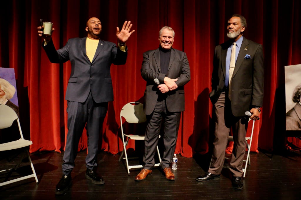 Three people stand onstage, a red curtain visible behind them.