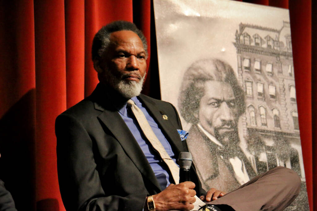 Kevin Douglass Greene is seated in front of a photo of his ancestor, Frederick Douglass.