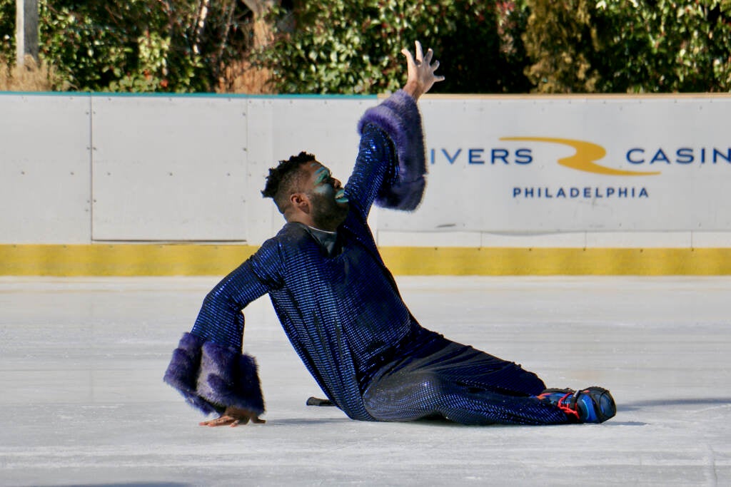 An ice skater in a blue outfit is sitting on the ice, reaching upward.