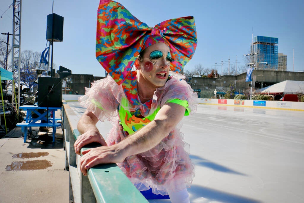 An ice skater and performer clutches the side of the ice skating rink on a sunny day.