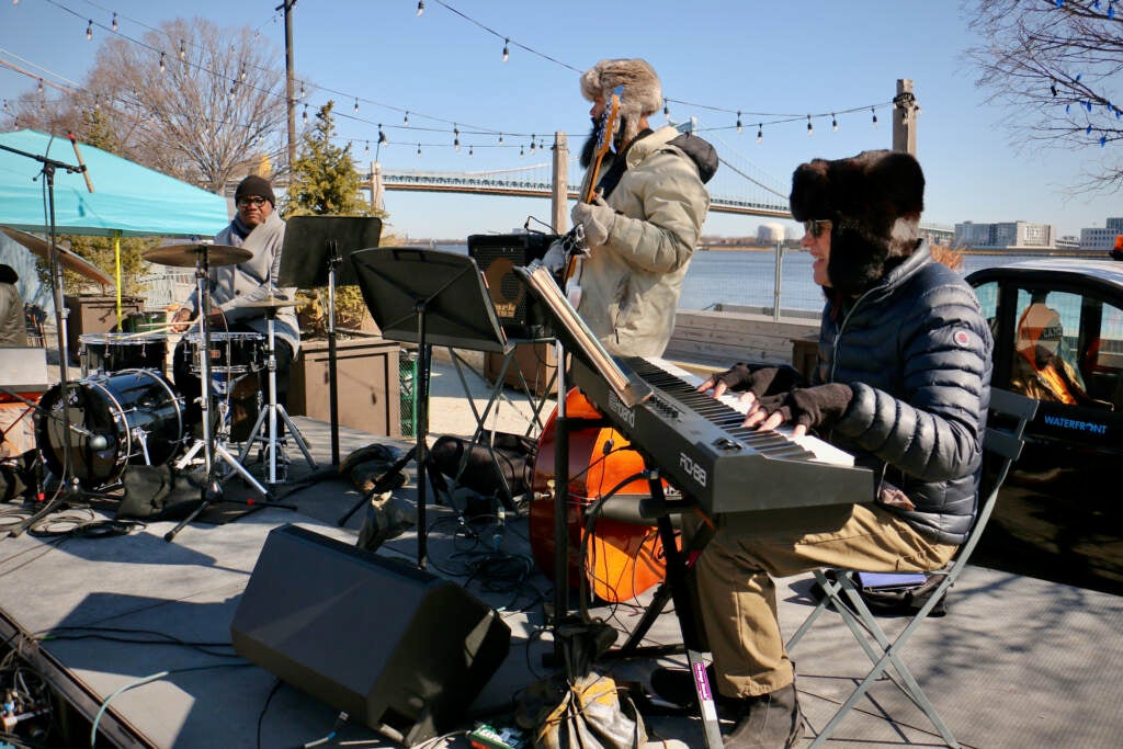 Musicians play the keyboard, drums and guitar on an outdoor stage.