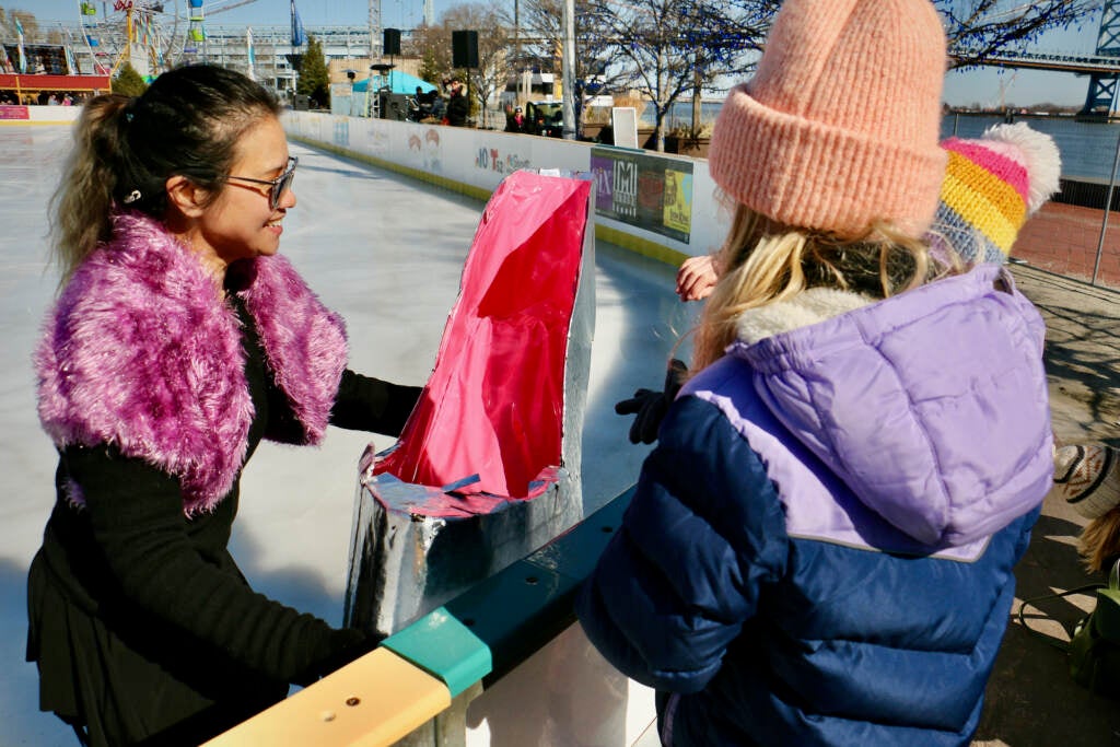 A performer and ice skater holds out a silver shoe for donations towards an observer on the other side of the rink.