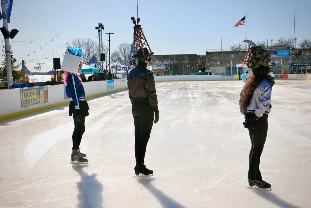 Three ice skaters are seen from behind as they stand in an empty ice-skating rink.