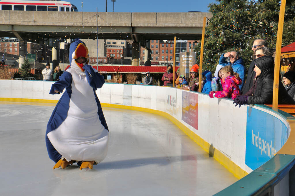 A penguin mascot skates by a crowd gathered on the side of the rink.