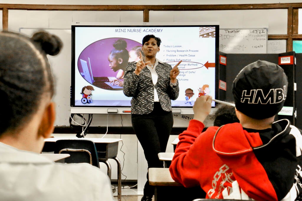 Angel McCullough stands at the front of a classroom. The backs of students' heads are visible in the foreground.