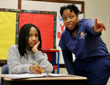 A nurse stands next to a student seated at the desk.