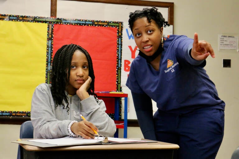 A nurse stands next to a student seated at the desk.