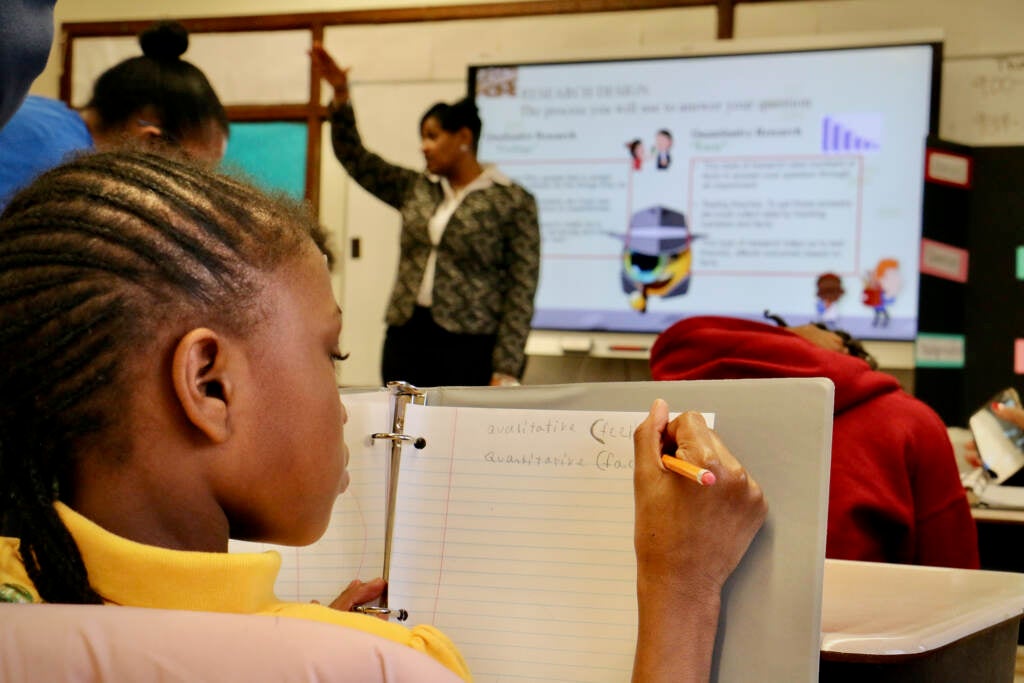 A student, seen from behind, writes in her notebook. In the background is a teacher demonstrating something at the front of the classroom.