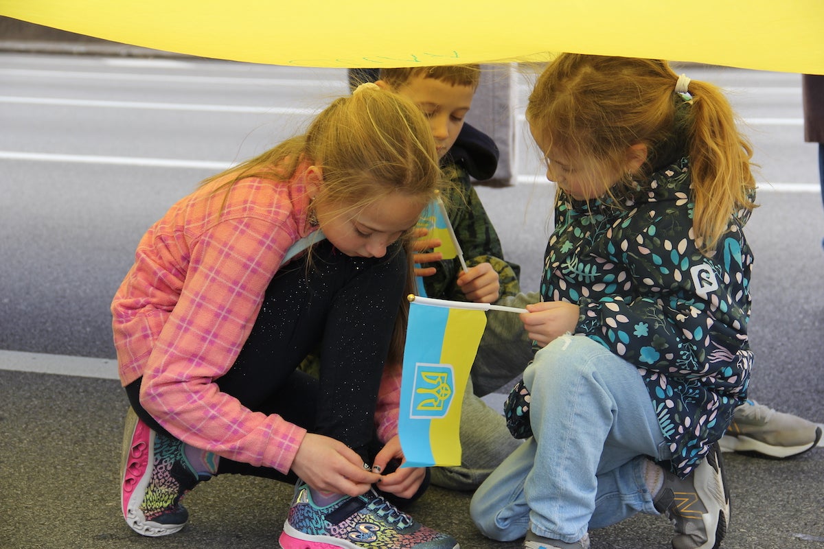 Three children huddle underneath a large flag of Ukraine