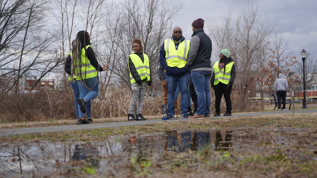 A group of people in yellow reflective vests talk together in a group on a pathway outdoors.