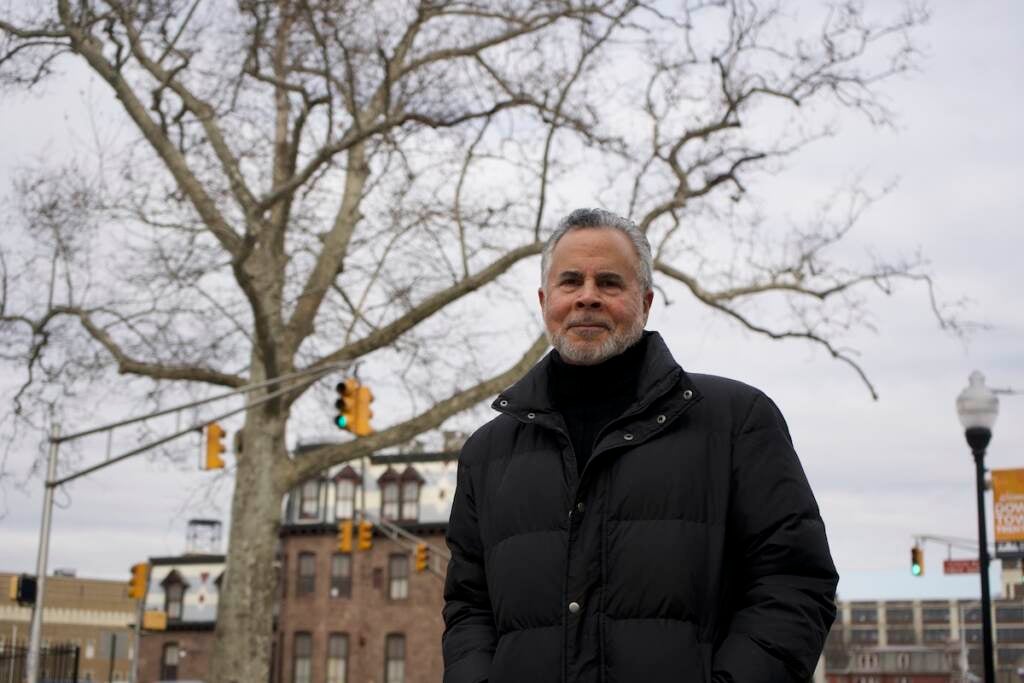 A man poses for the camera. A tree, buildings, and traffic lights are visible behind him.