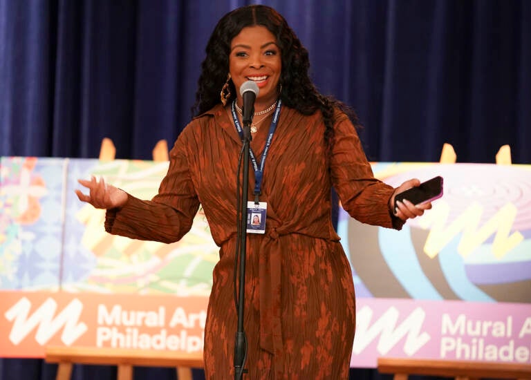 A woman speaks into a microphone onstage. Mural Arts logos are visible behind her.