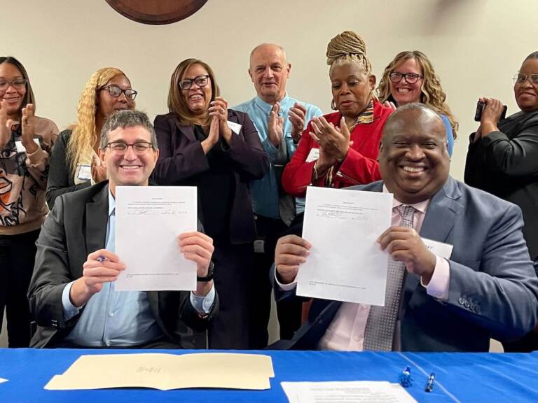 Two people hold up signed documents while seated at a table. Other people pose for a photo in the background.