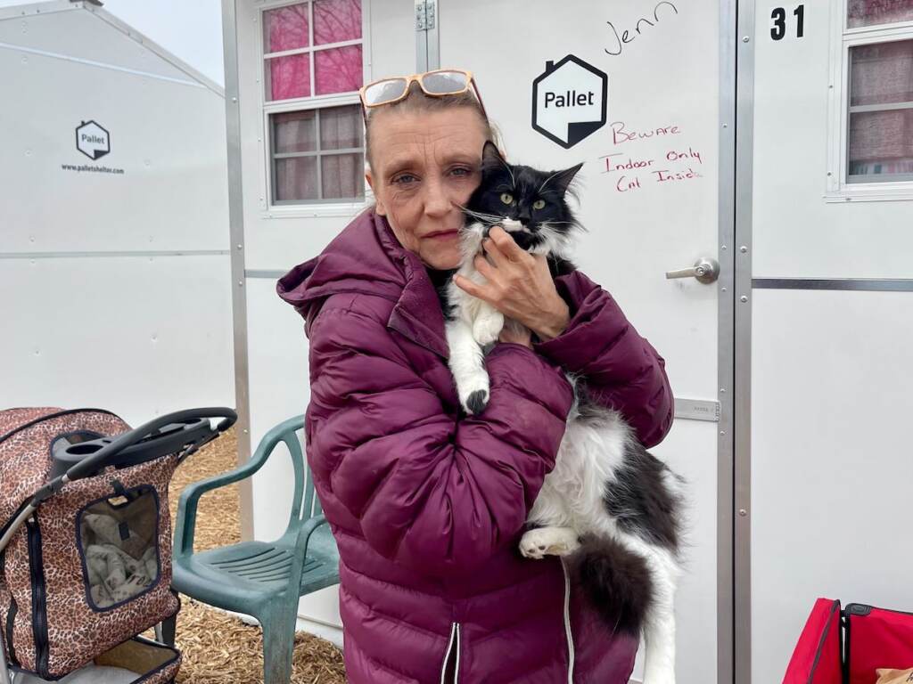 A woman holds up her cat outside of the white cabin she lives in at the Delaware Pallet Village.