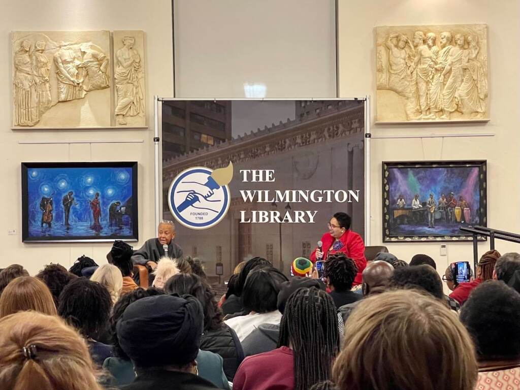 A stage is visible over the heads of a large audience gathered together at the Wilmington Public Library.