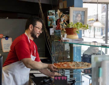 03 01 2023 k paynter Omar Quiñonez amy’s pizzeria-2 Owner Omar Quiñonez puts a pizza in the display case at Amy's Pizzeria. A customer is visible seated at a table on the other side of the counter.