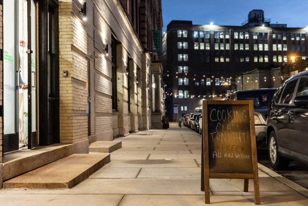 A sign on a sidewalk reads "Cooking Class Tonight! Free, All Are Welcome."