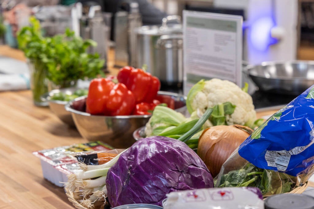 A close-up of fresh produce, including red bell peppers, a red onion, a yellow onion, a bag of spinach, and a cauliflower.