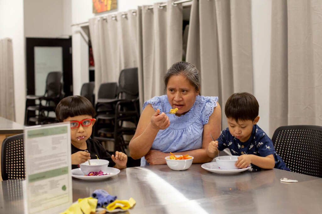 Felicitas Victoria, Joshua, and Lucas sit next to one another, eating a meal at a table.