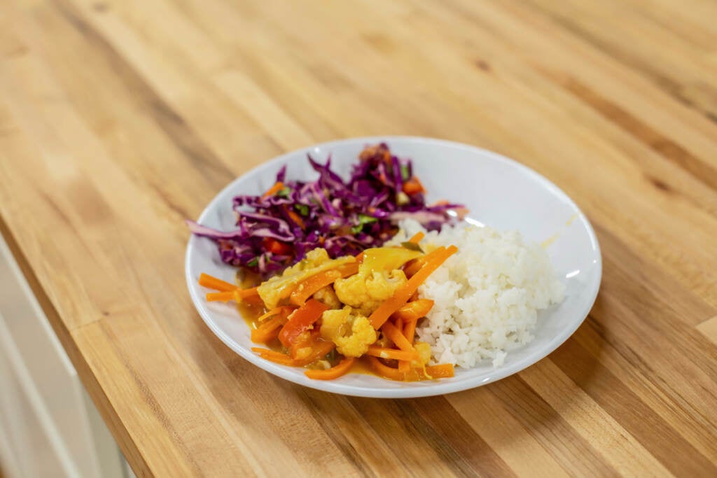 A close-up of a coconut Thai curry, a crunchy salad, and rice on a place placed on a wooden countertop.