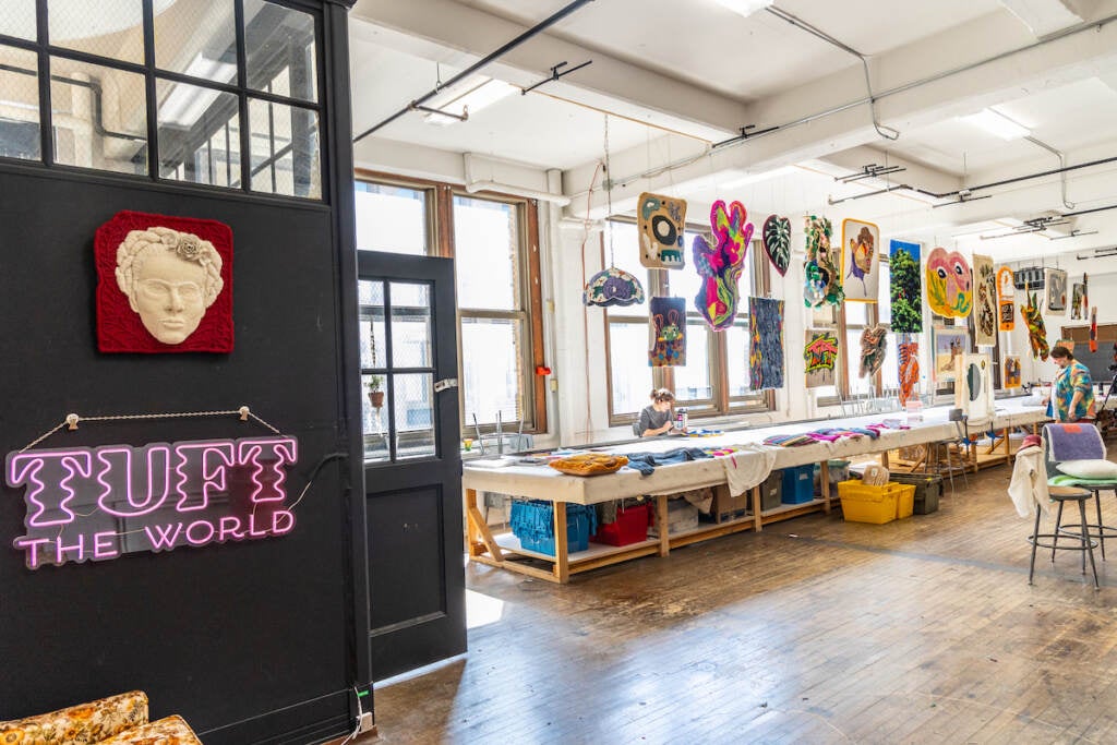 A light-filled room shows different tufted artworks, and a neon sign to the left reads "Tuft the World."