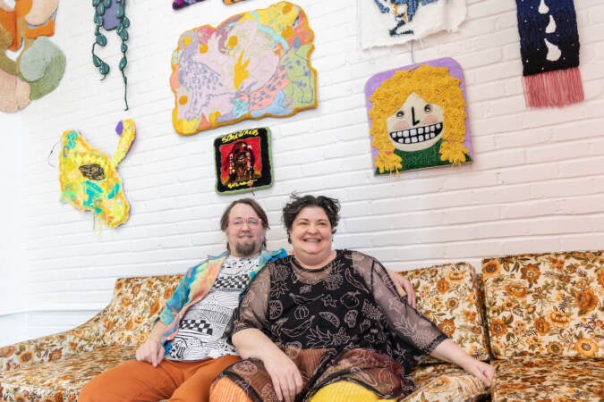 Tim Eads and Tiernan Alexander sit together on a sofa. Tufted artworks are hung on the wall above them.