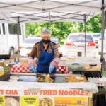 A vendor cooks on a grill at an open stand. Trees and cars are visible behind her.