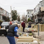 A person works on pieces of wood on a table in the street on a block of rowhomes.