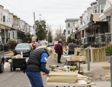 A person works on pieces of wood on a table in the street on a block of rowhomes.