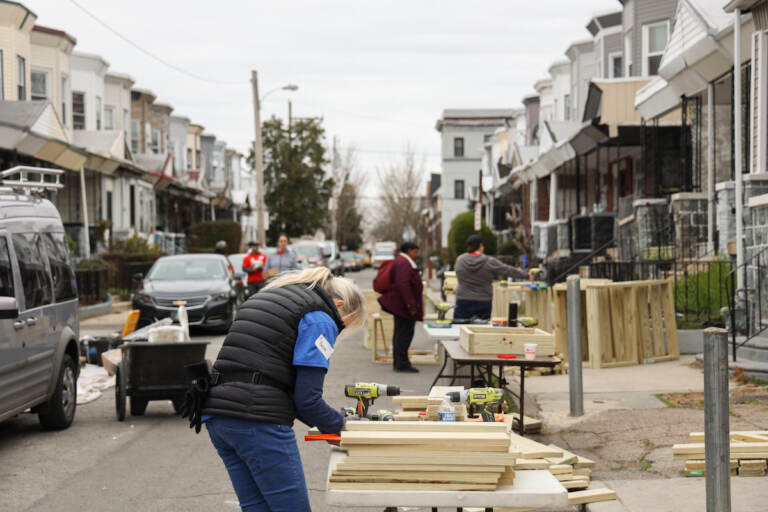 A person works on pieces of wood on a table in the street on a block of rowhomes.