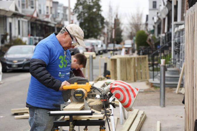 A person works on a piece of wood outdoors on a block of rowhomes.
