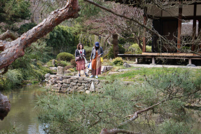 Two visitors walk next to a pond, near a wooden building. Tree branches are visible in the foreground.