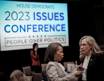 Two people talk in the foreground. A sign in the background reads: House Democrats 2023 Issues Conference.