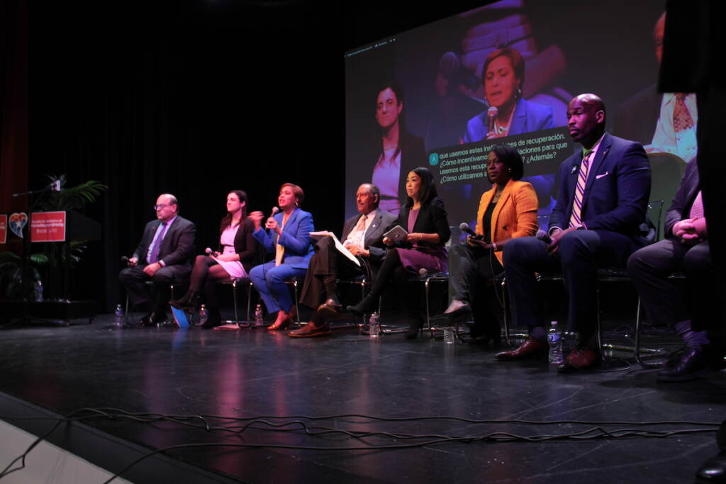 Mayoral candidates are seated in a row in chairs on a stage.