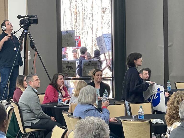 Protesters are visible through the windows of a meeting room.