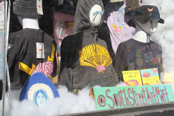 An egg design is displayed in a shore window where there are clothes hung on mannequins and a sign that reads @SouthStreetArtMart.