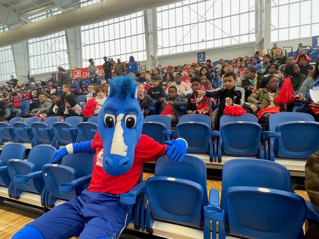 The Blue Coats' mascot Coaty, a blue horse, sits in the stands. Behind are numerous students and other members of the audience seated in the stands.