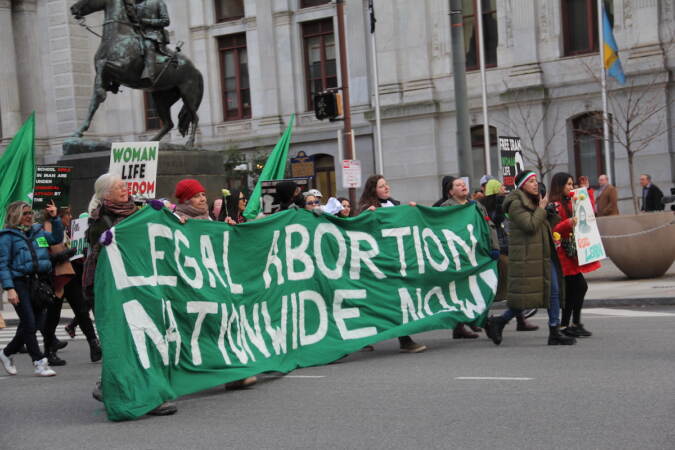 People march outside of City Hall holding a large green sign that reads 