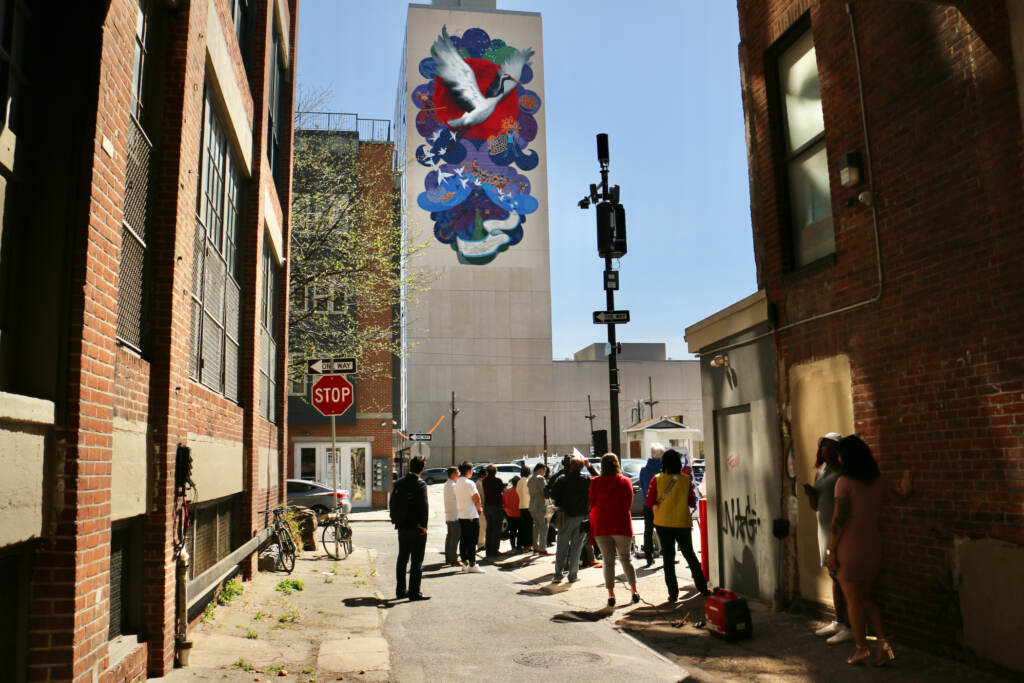 A crowd is visible on a street in front of a mural on a tall building.