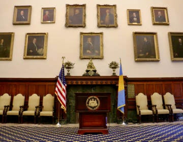 Portraits of former mayors line the walls of City Hall chambers. A podium and the U.S. flag and the Philadelphia flag are in the center of the photo. Chairs are lined up against the wall.