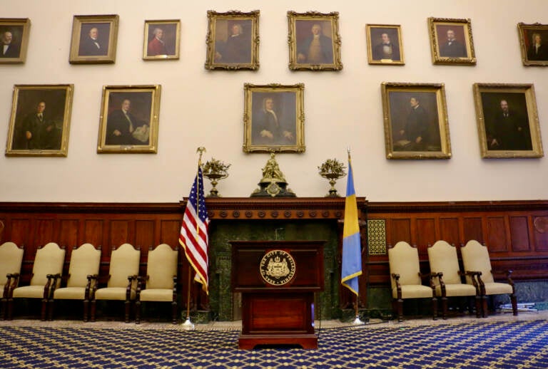 Portraits of former mayors line the walls of City Hall chambers. A podium and the U.S. flag and the Philadelphia flag are in the center of the photo. Chairs are lined up against the wall.