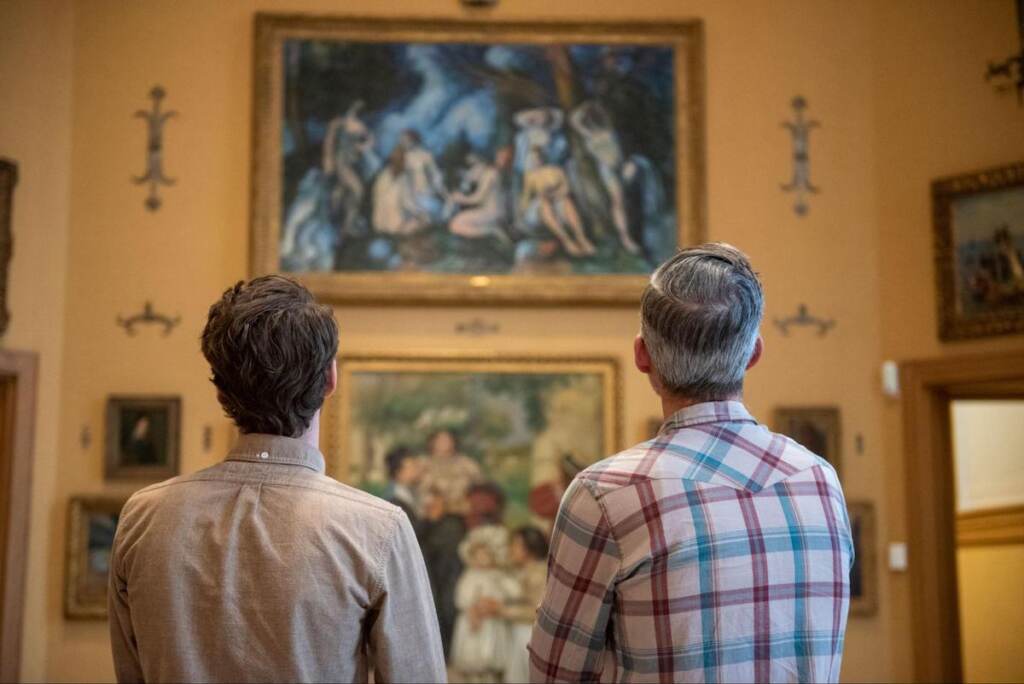 The backs of two people's heads are visible as they look at several paintings on the wall.