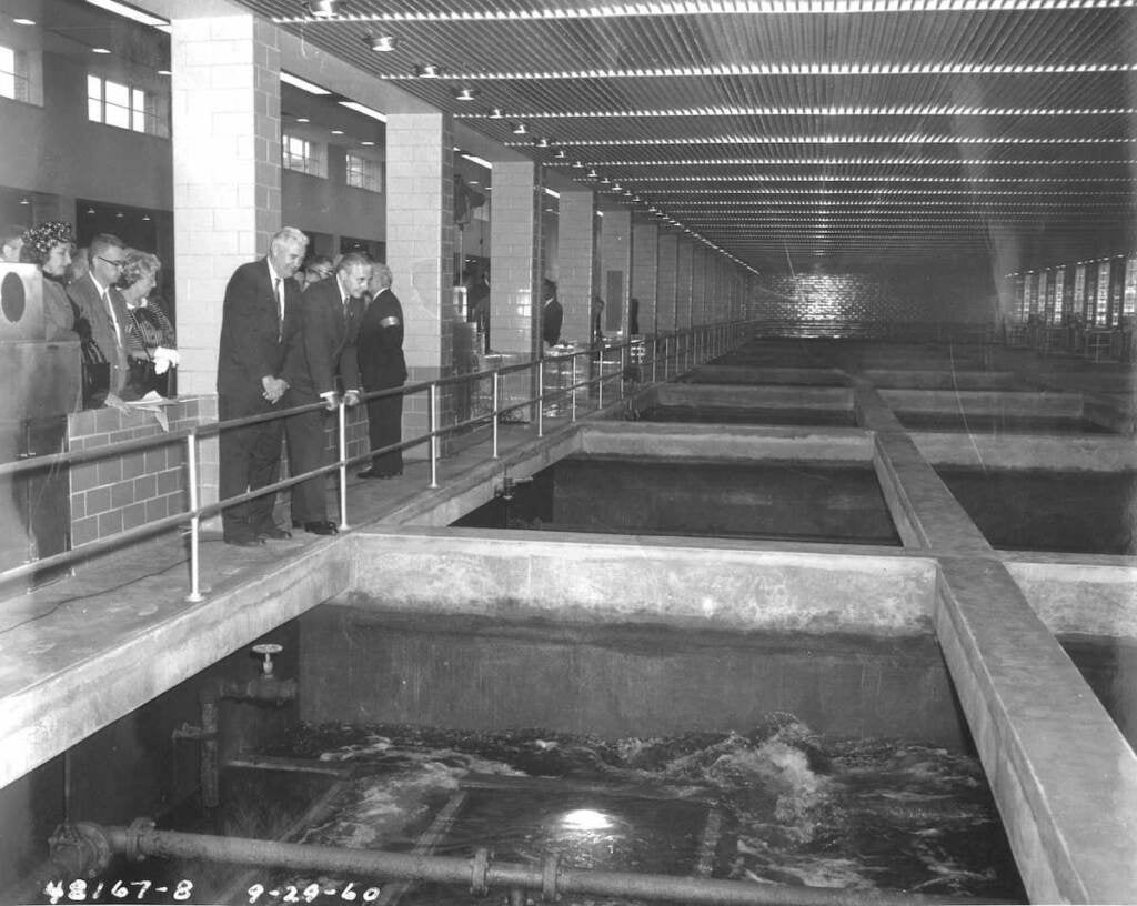A group of people overlook machinery and water in a water treatment plant in a black-and-white photo.