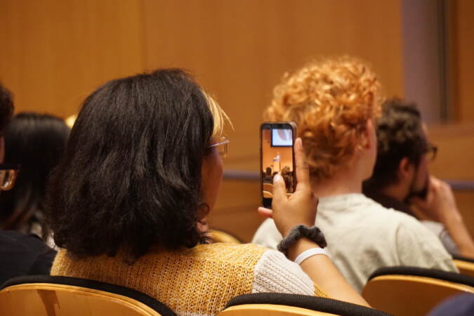 A student holds up a phone as they film a person speaking onstage.
