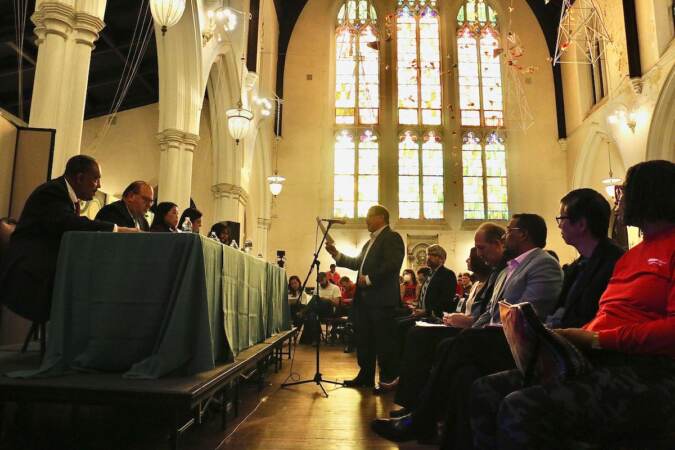 An audience is on one side of the photo, face-to-face with a table where a panel of candidates sit. The background shows a high, stained-glass church window.