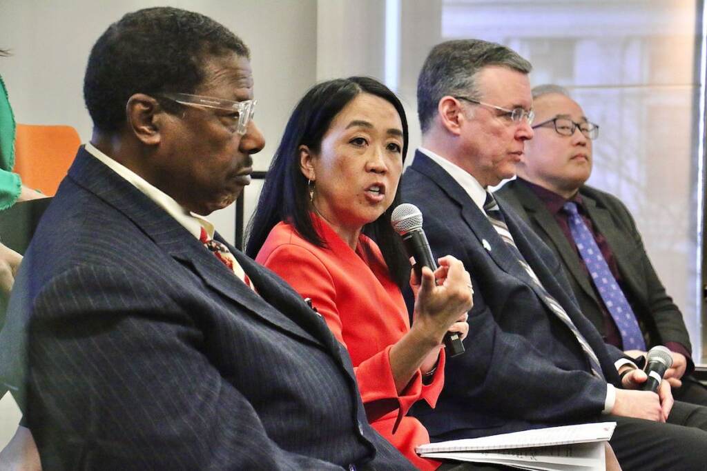 Helen Gym speaks into the microphone. Warren Bloom, Jeff Brown, and David Oh are seated nearby, listening.