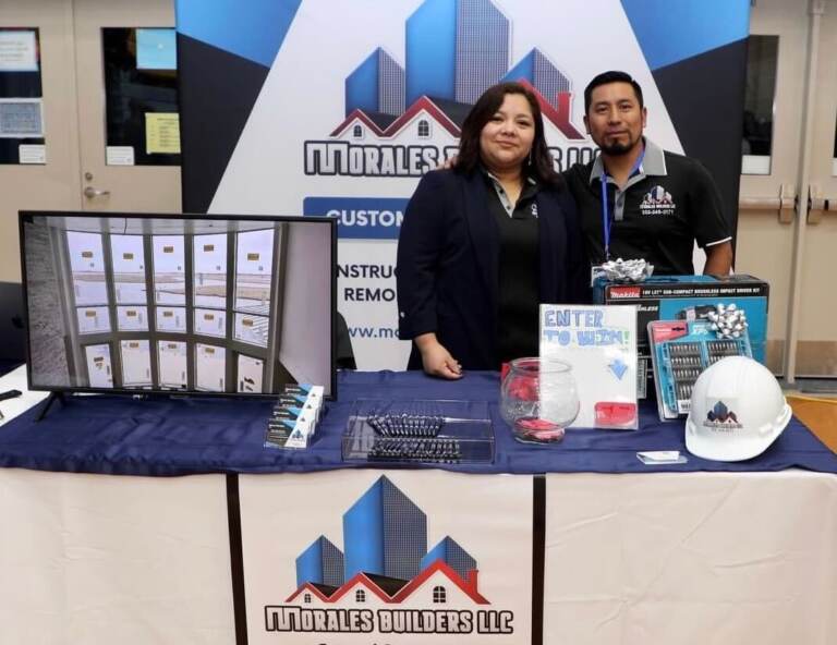 Sara Ovalle and Marco Morales pose for a photo, smiling, at a table advertising their business.