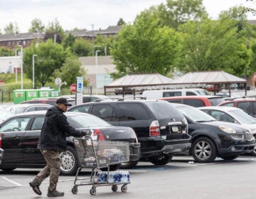 A person pushes a shopping cart in a parking lot. A case of bottled water is visible on the cart.