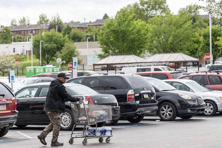 A person pushes a shopping cart in a parking lot. A case of bottled water is visible on the cart.