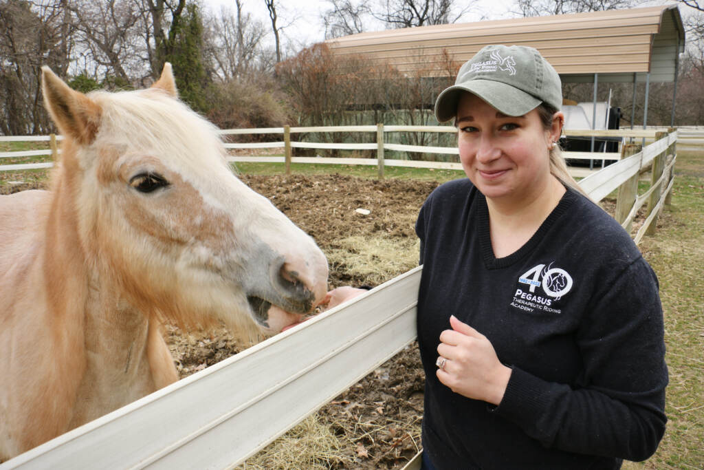 Teresa Fletcher looks towards the camera, feeding a horse that is next to her on the other side of a fence.